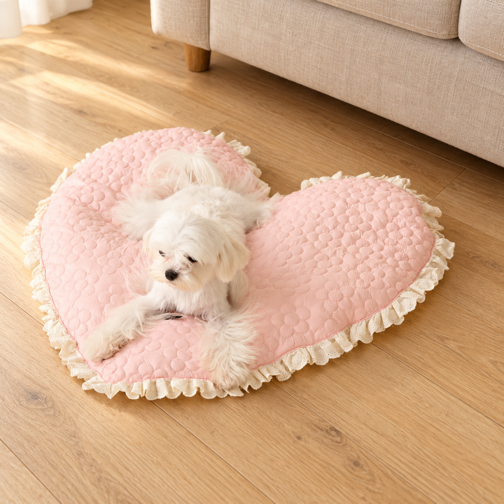 White dog resting on a Pink heart-shaped quilted pet mat with floral stitching and ivory ruffled trim in a warm home setting