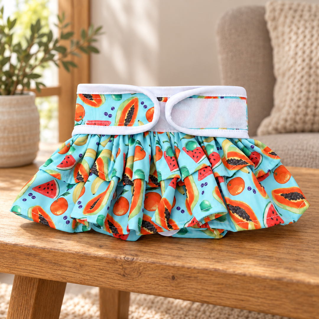Fruit-print ruffled pet diaper pants with white waist tabs and two gathered tiers on an oak home bench.