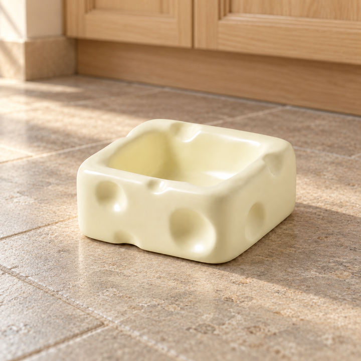 White small square cheese-shaped ceramic pet bowl on a natural stone kitchen floor.