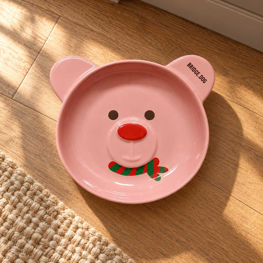 Pink BRIDGE.DOG Christmas Bear ceramic bowl on a warm oak floor beside a woven rug, retaining its red nose and red-and-green scarf design.