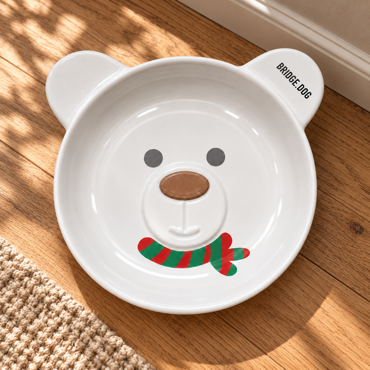 White BRIDGE.DOG Christmas Bear ceramic bowl with brown nose and red-and-green scarf design on a warm oak floor beside a woven rug.
