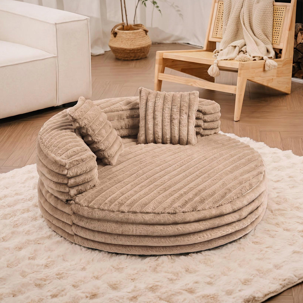 Khaki CloudCove round ribbed bolster bed with two pillows on a rug beside a wooden armchair