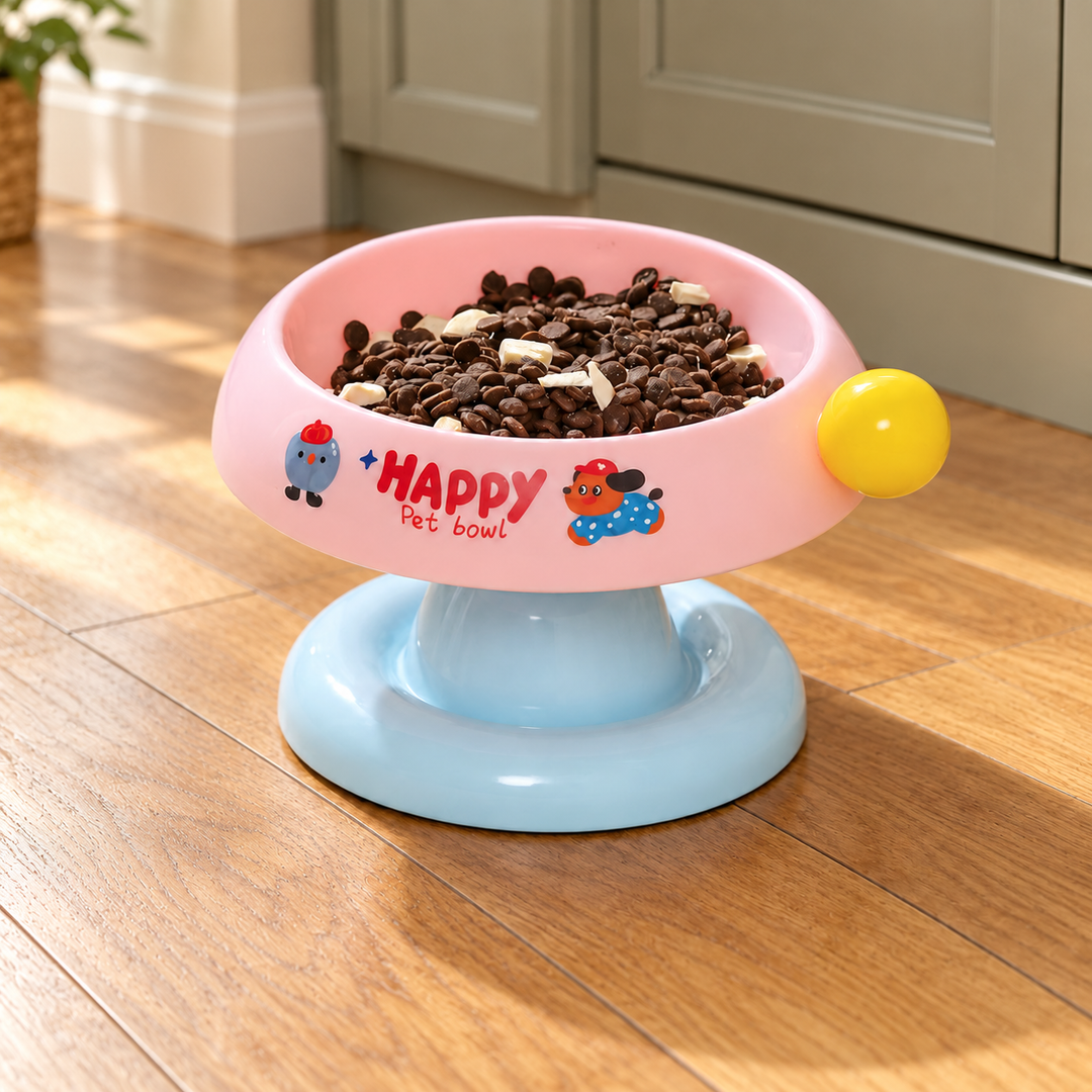 Pink tilted pet bowl with a light blue pedestal, yellow side ball and original illustrated print, shown in a home feeding corner.
