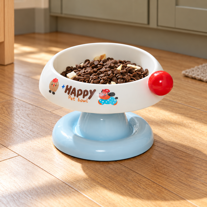White tilted pet bowl with a light blue pedestal, red side ball and original illustrated print, shown in a home feeding corner.