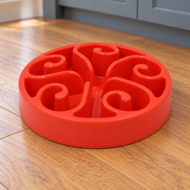 Red slow-feeder dog bowl with five curled spiral walls on a natural wood kitchen floor.